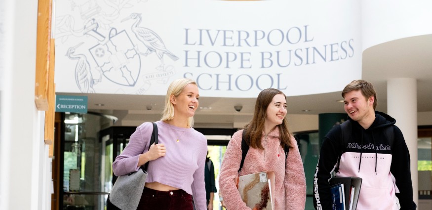 Two female students and one male student walk through a corridor with a Liverpool Hope Business School sign overhead on the wall behind them.